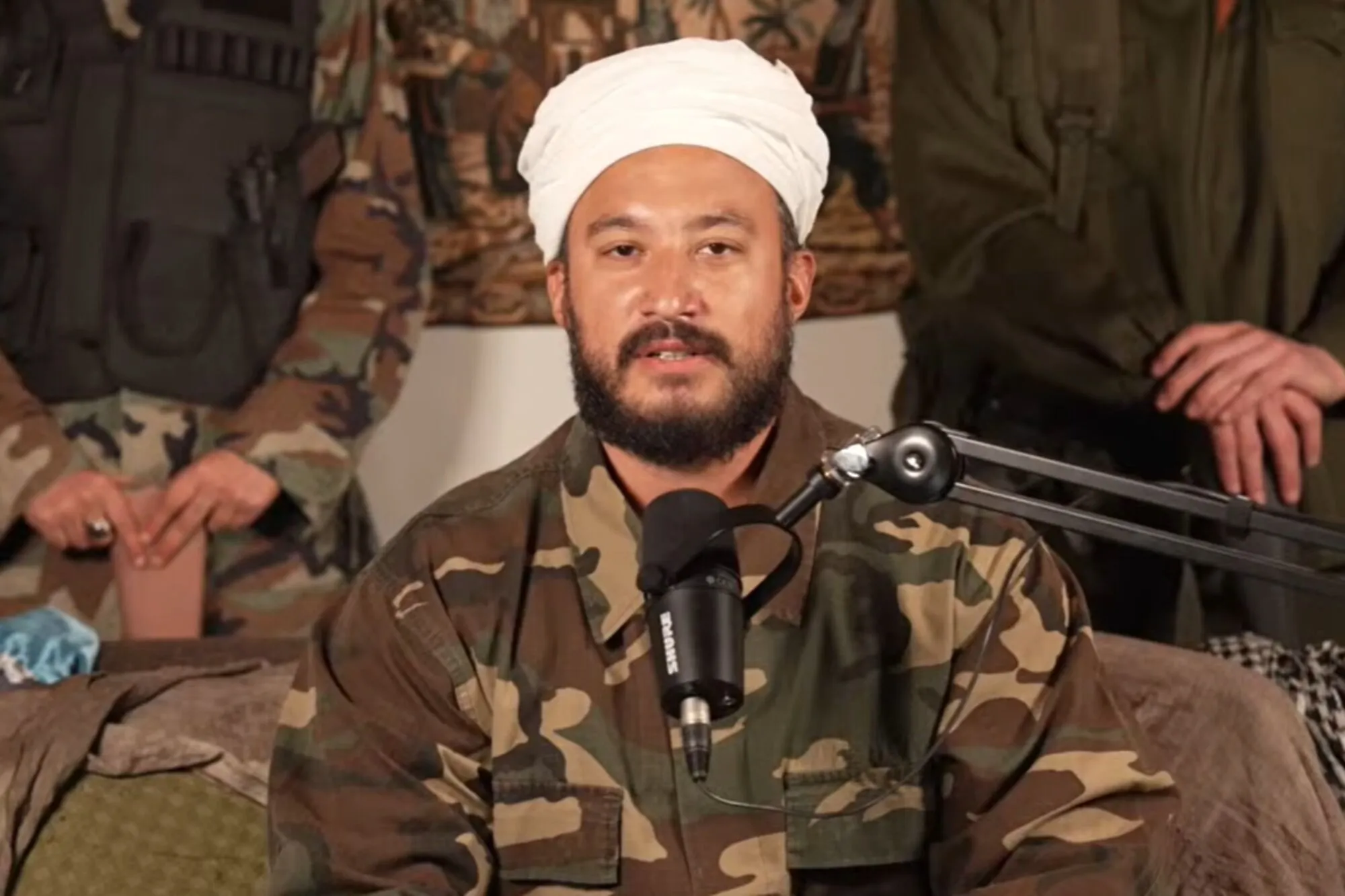 Tomás Rebord con turbante blanco y campera de camuflaje frente al micrófono